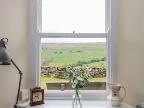 Ein Fenster mit Blick auf eine Steinmauer und Felder, eine Uhr, eine Vase mit Blumen und ein Teekessel auf einem Tisch.