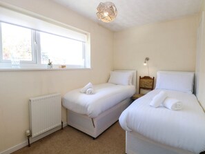 A bedroom with two beds, a window, a radiator, and a nightstand with a lamp. - Sweetacres Retreat in Hemsby (Great Yarmouth)