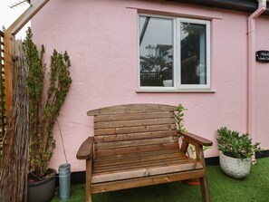 Un banc en bois se trouve devant un bâtiment rose, orné d’une fenêtre, d’une plante en pot et d’un baril en métal.