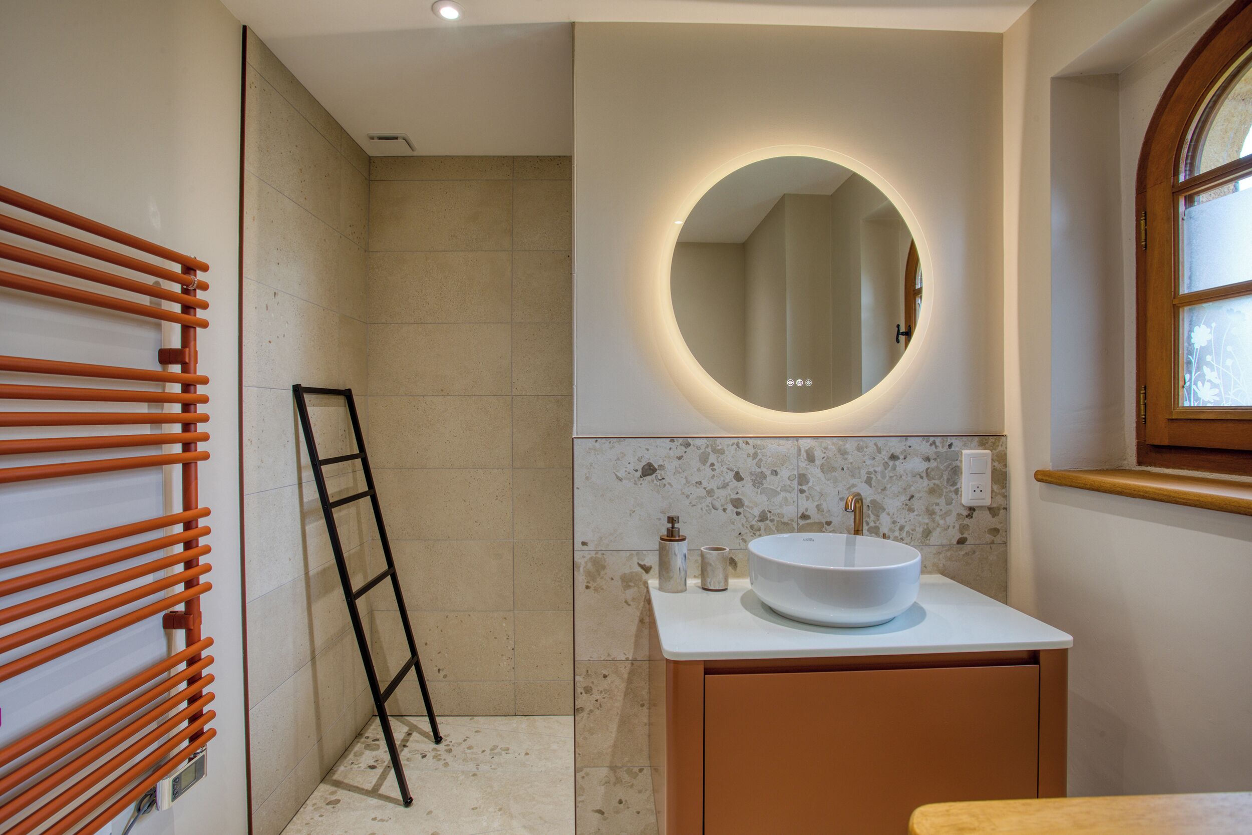 Une salle de bain avec un lavabo blanc, un miroir rond avec éclairage intégré, un porte-serviettes et un espace douche.