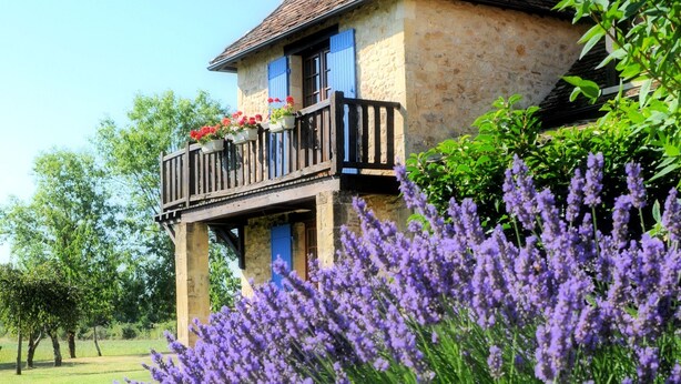 Un bâtiment en pierre doté d’un balcon, entouré d’une végétation luxuriante et de fleurs violettes.