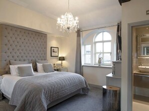 Un dormitorio con cabecera capitonada, candelabro, mesita de noche con lámpara, ventana con cortinas y baño con lavabo y espejo.