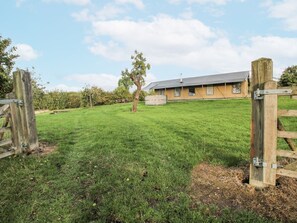 Ein grasbewachsenes Feld mit einem Holztor, einem Baum und einem Gebäude mit grauem Dach im Hintergrund.