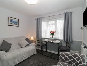 Ein Zimmer mit grauem Sofa, einem kleinen Tisch mit Blumenvase, einem Sessel und einem an der Wand montierten Fernseher.