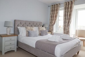 A neatly made bed with a grey headboard, a bedside table with a lamp, and a chair by the window. - 17 Ocean Heights is in Newquay (Newquay)