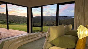 Une chambre dotée d’un grand lit, offrant une vue sur un paysage verdoyant grâce à de larges fenêtres, et donnant sur une terrasse en bois.