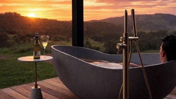 Un jacuzzi con vista a la puesta de sol y montañas, una botella de vino y una copa en una terraza de madera.
