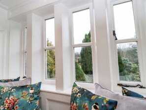 Ein Zimmer mit drei Fenstern, einer Bank mit floralen Kissen und Blick auf Grün.