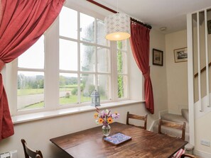 A dining area with a wooden table, chairs, a vase with flowers, a hanging light fixture, and a window with curtains. - Huntsman's Cottage, Cartmel (Grange-Over-Sands)