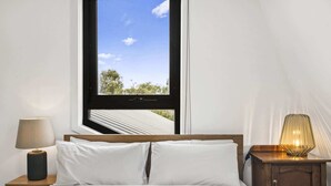A bedroom with a bed, a nightstand with a lamp, a wooden cabinet, and a window with a view of trees and a clear sky. - Comfortable 2 Bed Stay with Parking (Footscray)