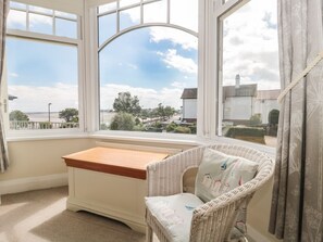 Ein Zimmer mit einem großen Erkerfenster, einem Korbstuhl, einem kleinen Holztisch und Blick auf einen Fluss und Gebäude.