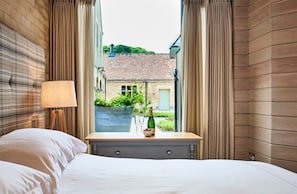 Habitación de hotel con cama, ventana con cortinas, mesita de noche con una botella y vasos, y vistas al exterior.