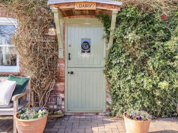 Una puerta verde con un letrero que dice 'DAIRY' encima, un muro de ladrillo y una planta en maceta al frente.