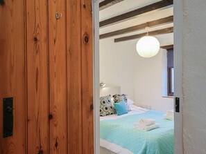 Eine Holztür führt in ein Schlafzimmer mit einem Bett, Kissen, einer Pendelleuchte und einem Fenster.