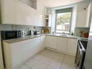 A kitchen with white cabinets, a microwave, a toaster, a sink, and a window with a view of greenery outside. - Trwst Y Don, Rhosneigr (Rhosneigr)