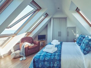 Ein Schlafzimmer mit schrägverdeckter Decke, einem Bett mit gemustertem Überwurf, einem braunen Sessel, einem Fernseher auf einem kleinen Tisch und einem Kleiderschrank.