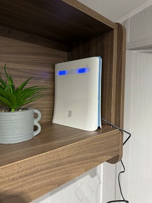 Un appareil blanc avec une lumière bleue, posé sur une étagère en bois, une tasse blanche avec une plante, et un câble noir.
