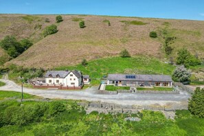 A rural setting with two main buildings, one with a white facade and another with a grey roof, surrounded by greenery and a hill in the background. - Charming Maesgwyn Retreat in Beguildy (Knighton)