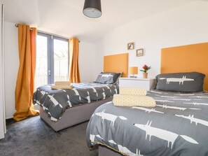 A bedroom with two beds, a window with curtains, a bedside table with a lamp, and a wall-mounted shelf with pictures.