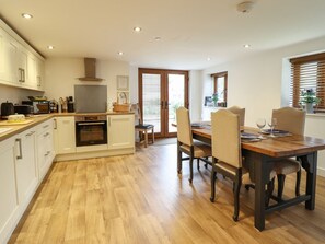 A modern kitchen with white cabinets, a built-in oven, and a dining area with a wooden table and chairs. - The Granary, Llandyssil, Powys (Montgomery)