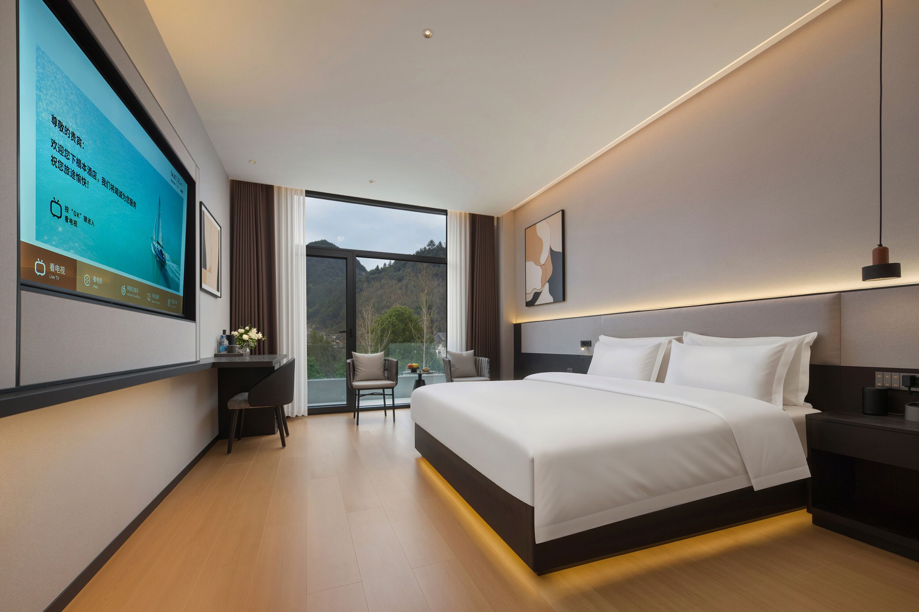 A modern hotel room with a large bed, a desk, and a TV screen displaying a scenic view.