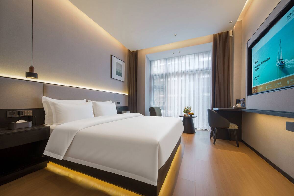 Quarto de hotel moderno com uma cama grande, uma escrivaninha, televisão e uma janela ampla com cortinas.