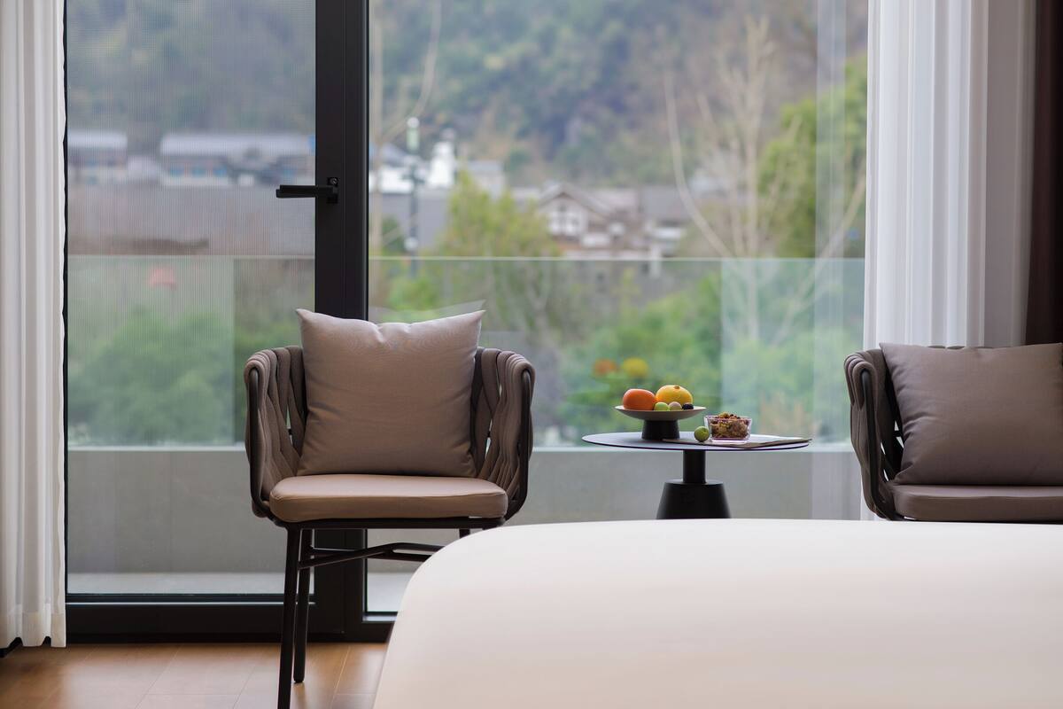 Quarto de hotel moderno com uma janela ampla, duas poltronas, uma mesa com frutas e uma cama.