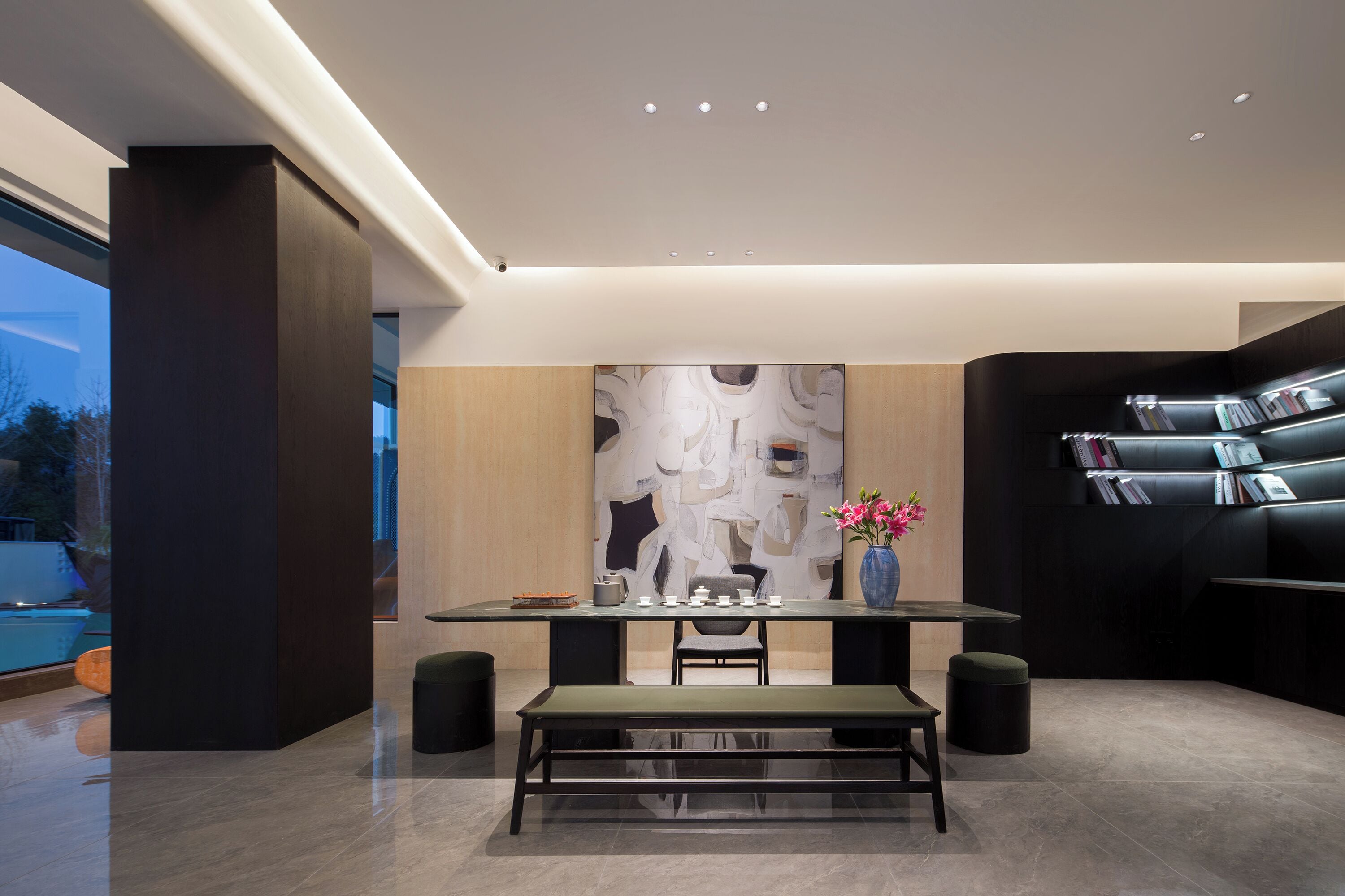 Un espace repas moderne avec une table en verre, des chaises noires et un banc. Un grand tableau abstrait orne le mur, et une bibliothèque contient des livres et des magazines.