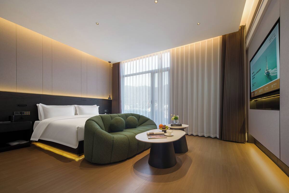 Quarto de hotel moderno com cama, sofá verde, uma mesinha com livros e televisão na parede.