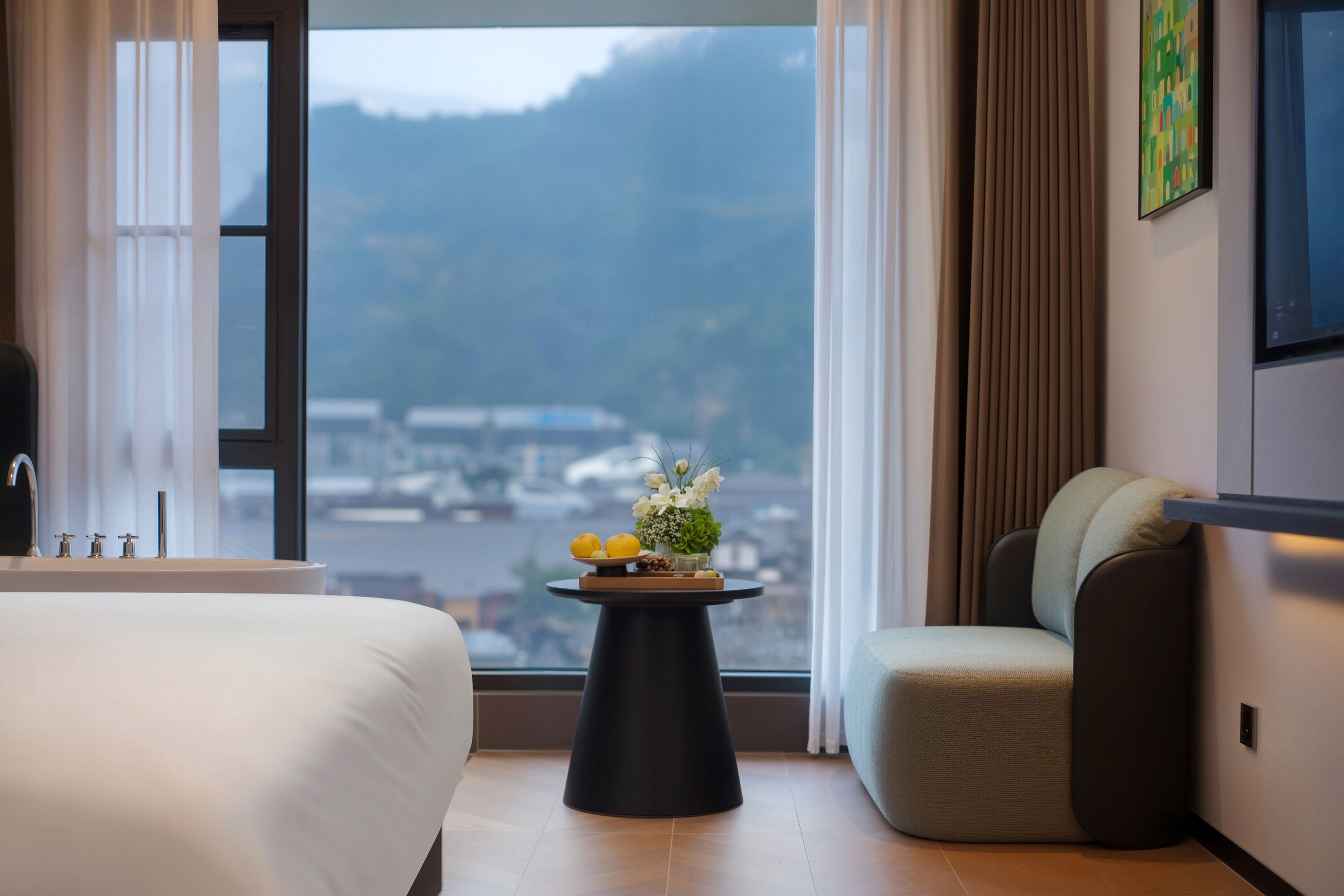 Habitación de hotel con cama, una mesita redonda con un arreglo floral, una silla y vista a un paisaje urbano a través de ventanas amplias.