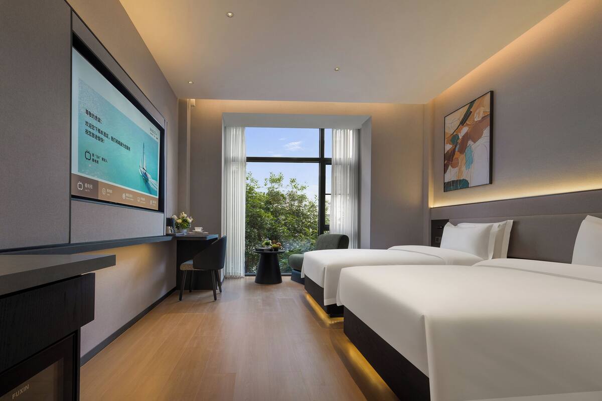 Quarto de hotel moderno com uma TV grande, duas camas, uma escrivaninha e vista para o verde pela janela.