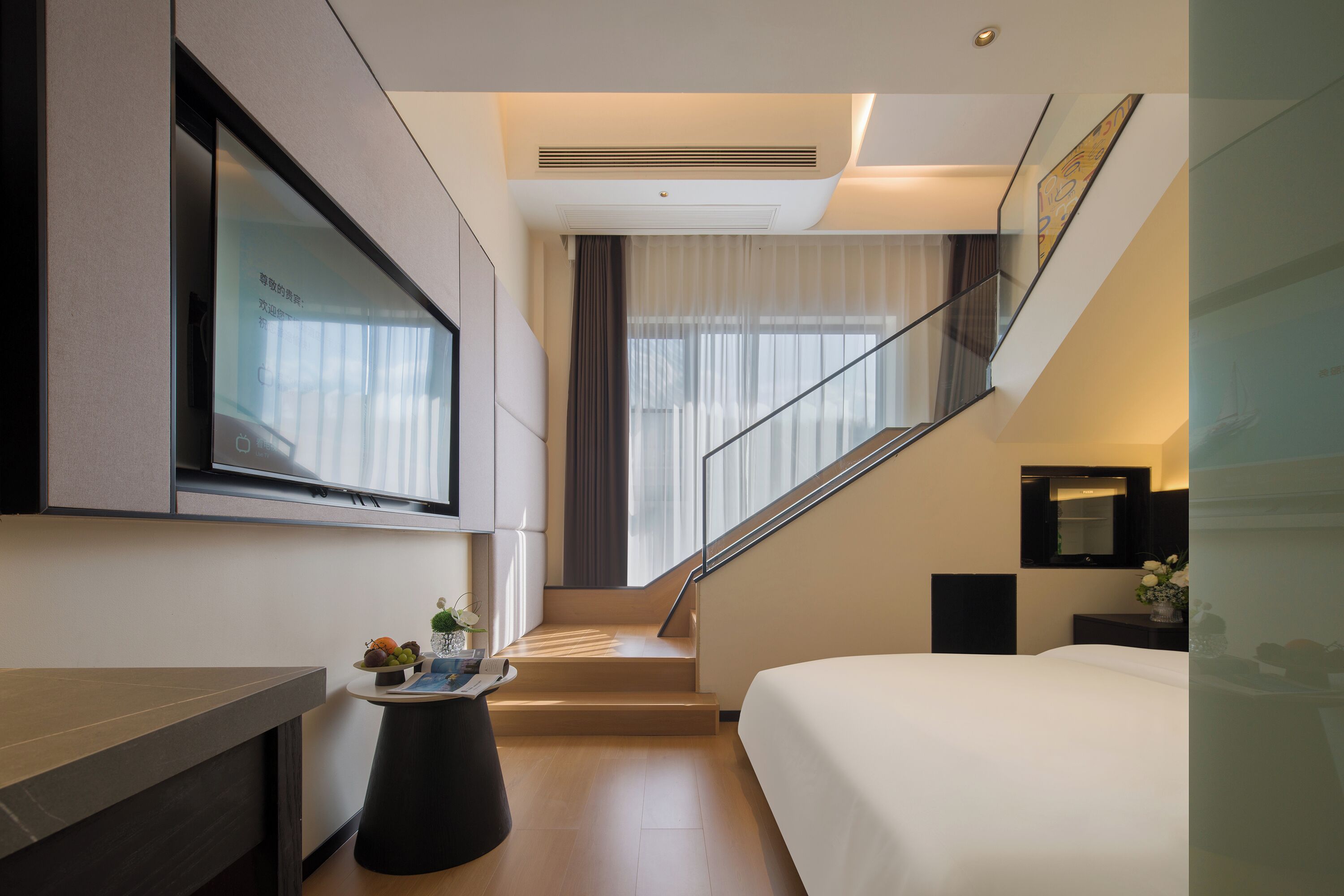 A modern hotel room with a bed, a TV, a small table with fruits, a staircase, and a window with curtains.