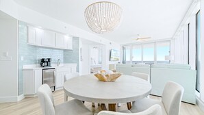 A modern kitchen with white cabinets, a dining area with a round table, and a view of the sea through large windows. - Colonnades 1403 | Completely updated! Gorgeous condo, beachfront, sleeps 12! (Gulf Shores)