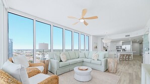 A spacious living area with a large window, a ceiling fan, a light-colored sofa, and a kitchen area in the background. - Colonnades 1403 | Completely updated! Gorgeous condo, beachfront, sleeps 12! (Gulf Shores)