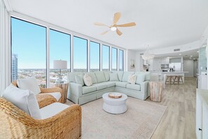 Un vaste espace de vie avec une grande fenêtre, un ventilateur de plafond, un canapé de couleur claire et un coin cuisine à l’arrière-plan.