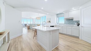 A modern kitchen with white cabinetry, a large island, and a dining area with a view. - Colonnades 1403 | Completely updated! Gorgeous condo, beachfront, sleeps 12! (Gulf Shores)