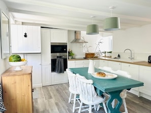 Une cuisine moderne avec des meubles blancs, une table à manger de couleur turquoise, des chaises blanches, un buffet en bois et un luminaire suspendu.