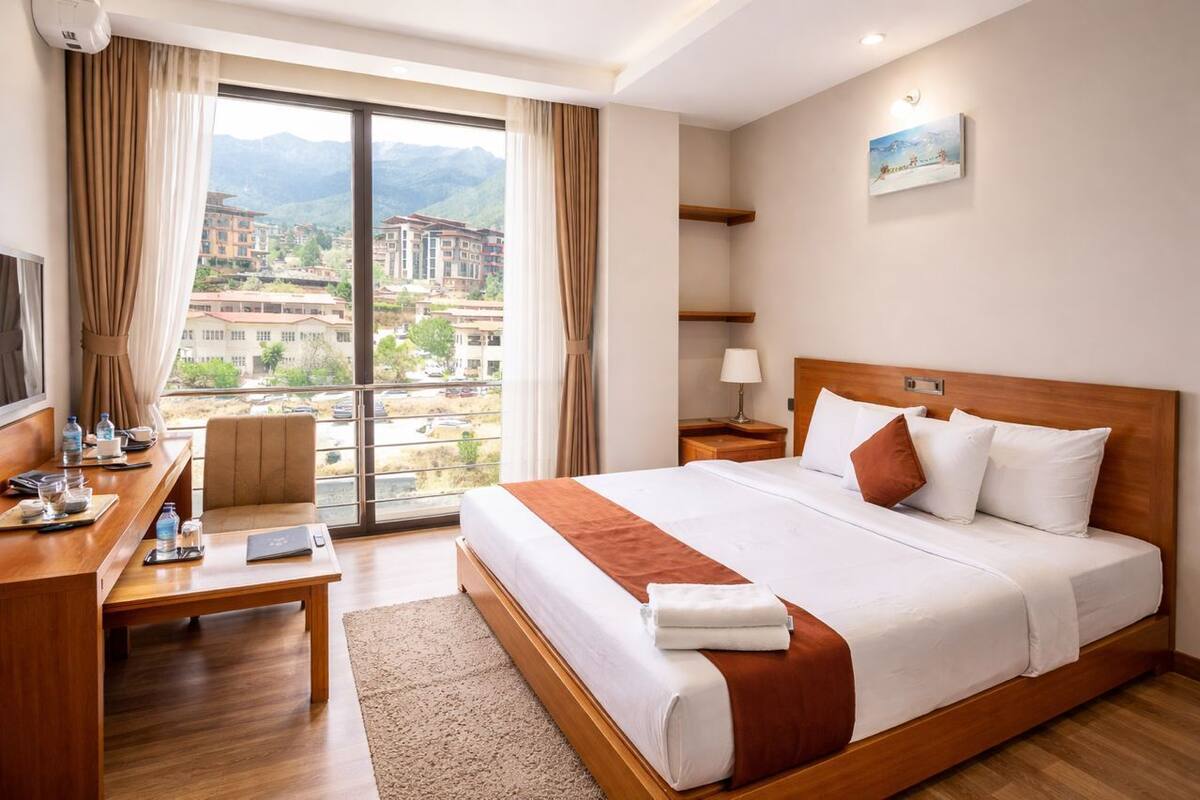 A hotel room with a bed, a desk, a chair, a lamp, and a view of buildings and mountains.