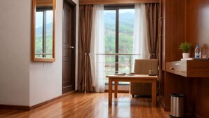A modern room with wooden flooring, a large window with curtains, a small table, and a mirror on the wall. - Hotel HTC Thimphu (Thimphu)