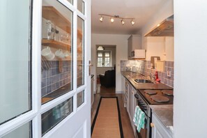A kitchen with a stove, sink, and cabinets, a window with dishes, and a hallway leading to a living area. - Kestrel Cottage, Bassenthwaite (Keswick)