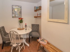 A dining area with a round table, two grey chairs, a white vase with flowers, a wine bottle, and a small book on a wooden side table. - Kestrel Cottage, Bassenthwaite (Keswick)