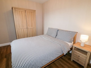 A bedroom with a bed, checkered bedding, a nightstand with a lamp, and a wooden wardrobe.
