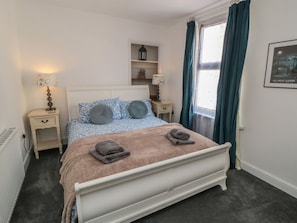 A neatly made bed with a headboard, two bedside tables with lamps, a shelf with decorative items, and a window with curtains. - 1 Albion Place, Whitby (Whitby)