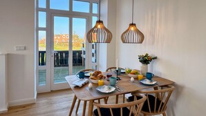 Ein Essbereich mit einem für das Frühstück gedeckten Tisch, Holzstühlen, hängenden Pendelleuchten und Blick durch eine Glastür.