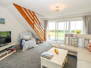 A living room with a TV, books, a coffee table, a sofa, a chair, and a staircase. - Gull Cottage, Kessingland (Lowestoft)