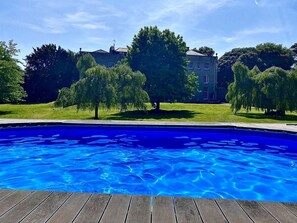 A swimming pool with a wooden deck, surrounded by trees and a building in the background. - Frenchay Park House, Bristol (Bristol)