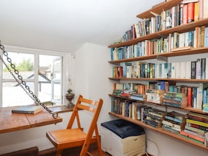 Ein Zimmer mit Schreibtisch aus Holz, einem Stuhl, einer Bücherwand voller Bücher und einem Fenster mit Blick nach draußen.