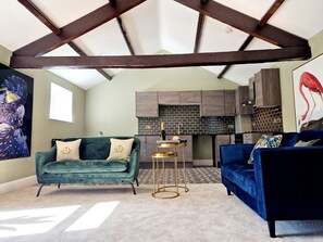 A loft-style living area with a high ceiling, exposed beams, a fireplace, and a kitchen in the background.