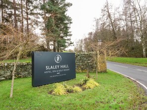 A sign for Slaley Hall Hotel, Spa & Golf Resort, part of The QHotels Collection, with a stone wall and trees in the background. - Slaley Park Villa, Hexham (Hexham)