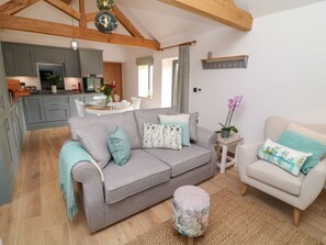 A cozy living room with a grey sofa, a smaller white sofa, and a round ottoman. A kitchen is visible in the background with grey cabinets and a dining table. - Tawny Barn, Woodhall Spa (Lincoln)